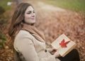 Side view of peaceful young female in warm coat and scarf sitting on bench with open book in hands and looking away pensively while resting in autumn park
