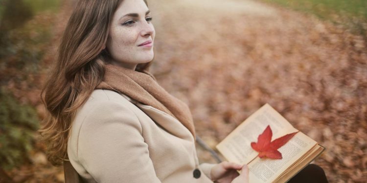 Side view of peaceful young female in warm coat and scarf sitting on bench with open book in hands and looking away pensively while resting in autumn park