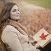 Side view of peaceful young female in warm coat and scarf sitting on bench with open book in hands and looking away pensively while resting in autumn park