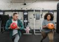 Young muscular ethnic male instructor in activewear doing lunges exercise with ball near plus size black woman during training in sport club