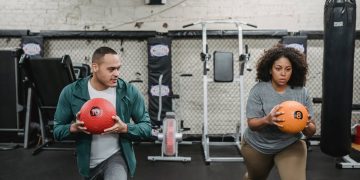 Young muscular ethnic male instructor in activewear doing lunges exercise with ball near plus size black woman during training in sport club