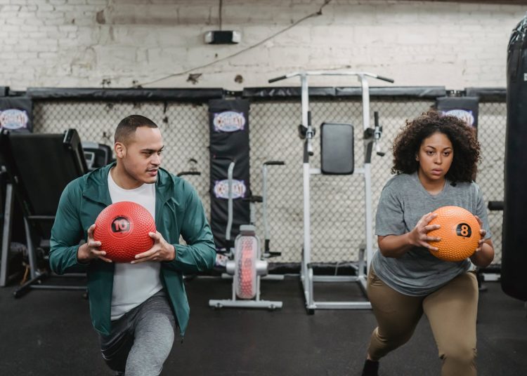 Young muscular ethnic male instructor in activewear doing lunges exercise with ball near plus size black woman during training in sport club