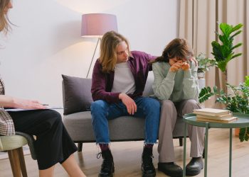 Young male embracing frustrated girlfriend during session with psychologist in light room in daytime