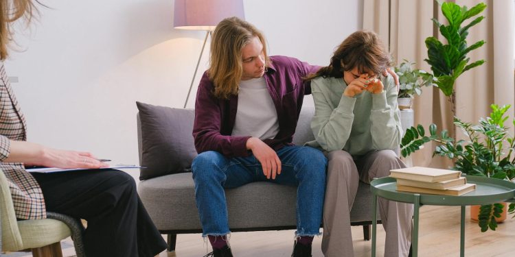 Young male embracing frustrated girlfriend during session with psychologist in light room in daytime